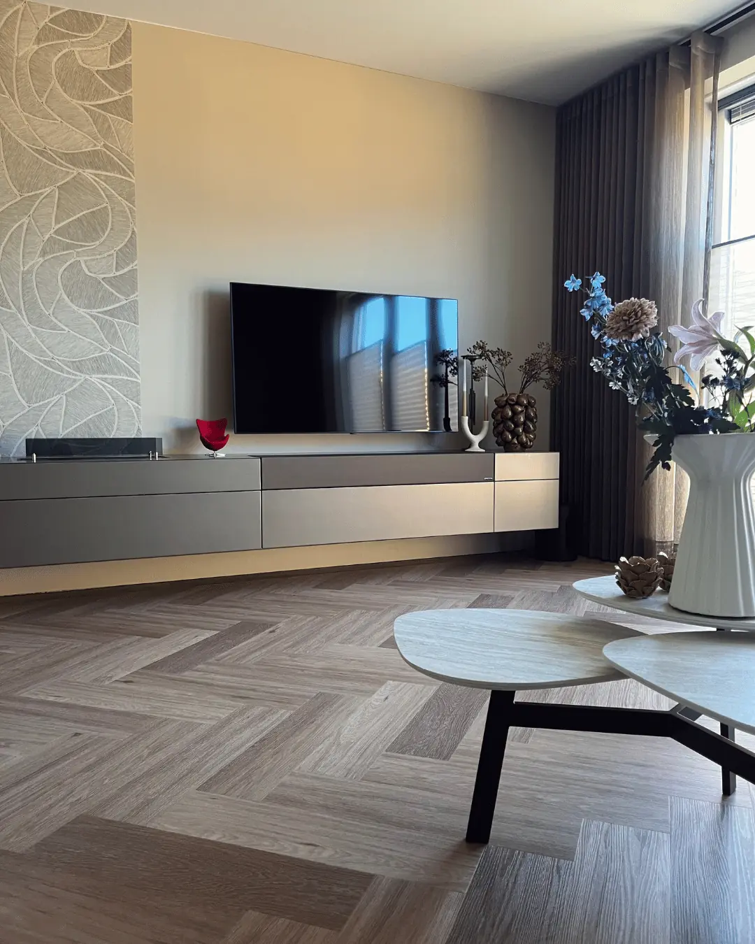 Woonkamer met zwevend dressoir, tv-wand, bloemen en ronde salontafels op een visgraatvloer