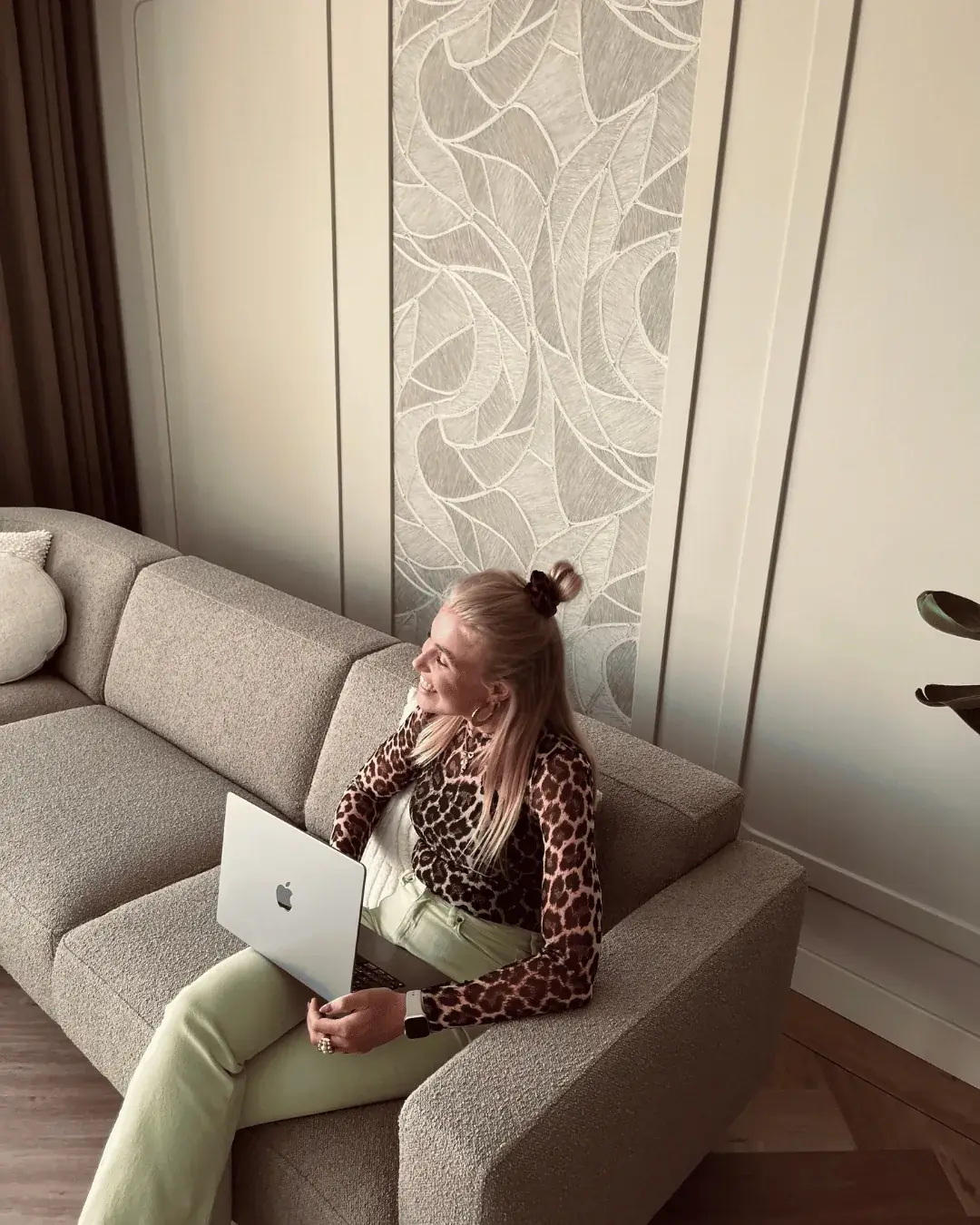Trijntje zit met laptop op de grijze hoekbank voor het omlijste behangpaneel in de woonkamer