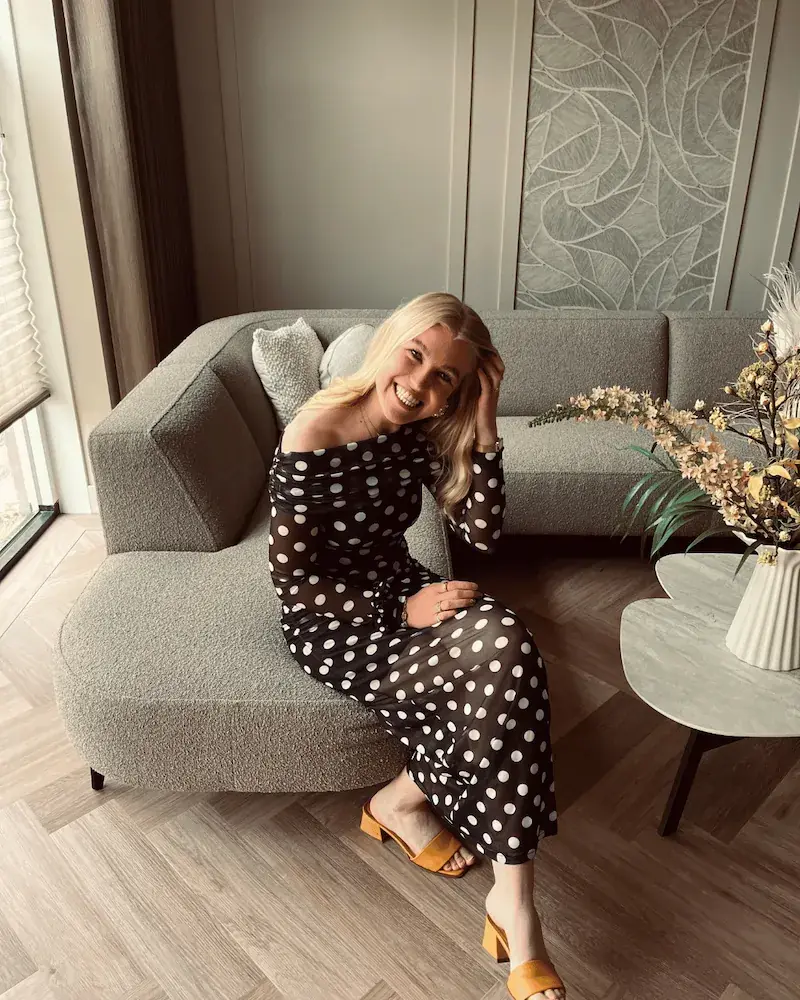 Trijntje Laan zit glimlachend op de afgeronde hoek van een grijze bouclé bank met haar hand in haar haar, op de salontafel een witte ribbelvaas met bloemen